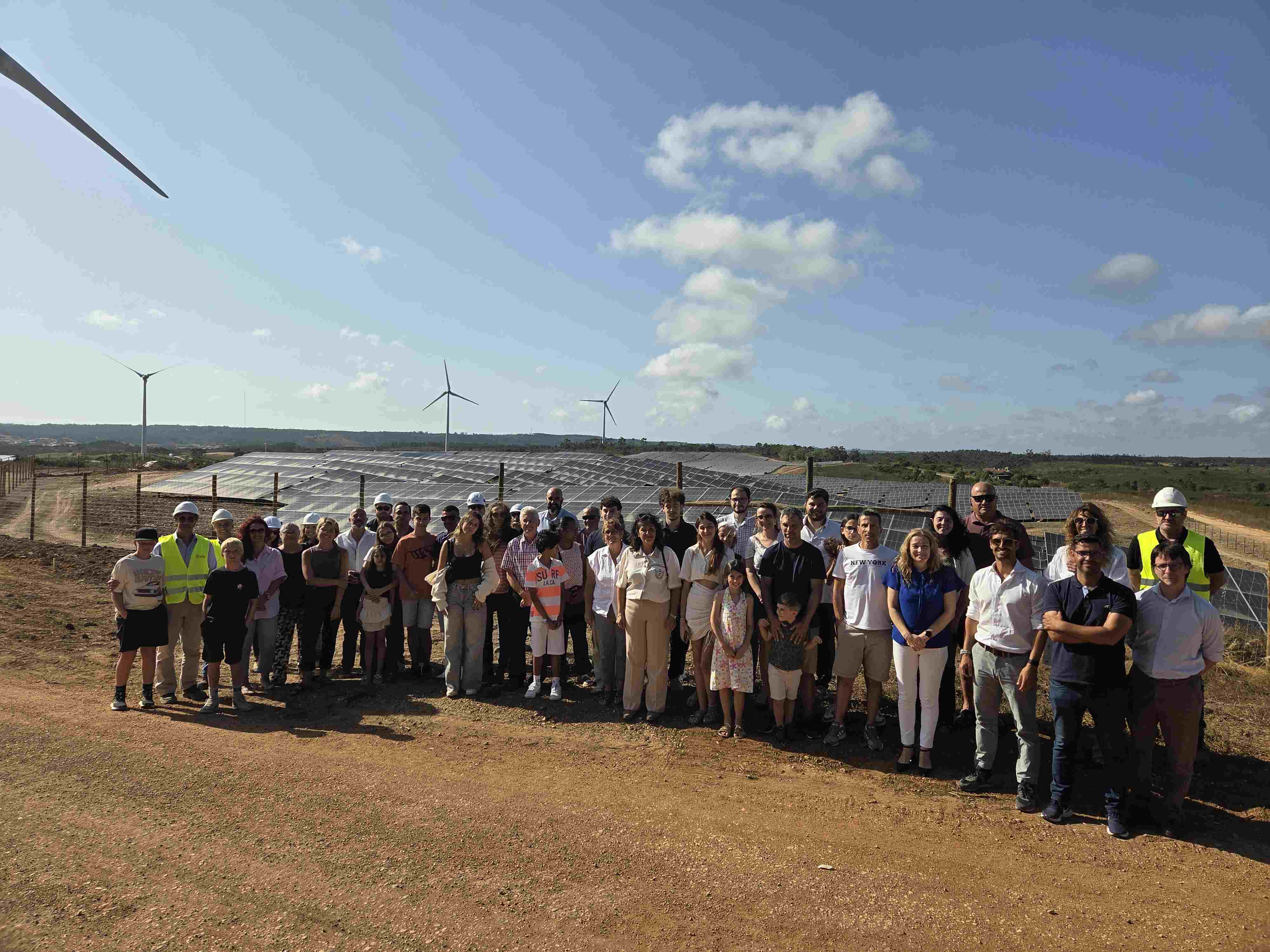 Grupo de pessoas posando para uma foto em frente a um parque de instalação solar e alguns moinhos de vento