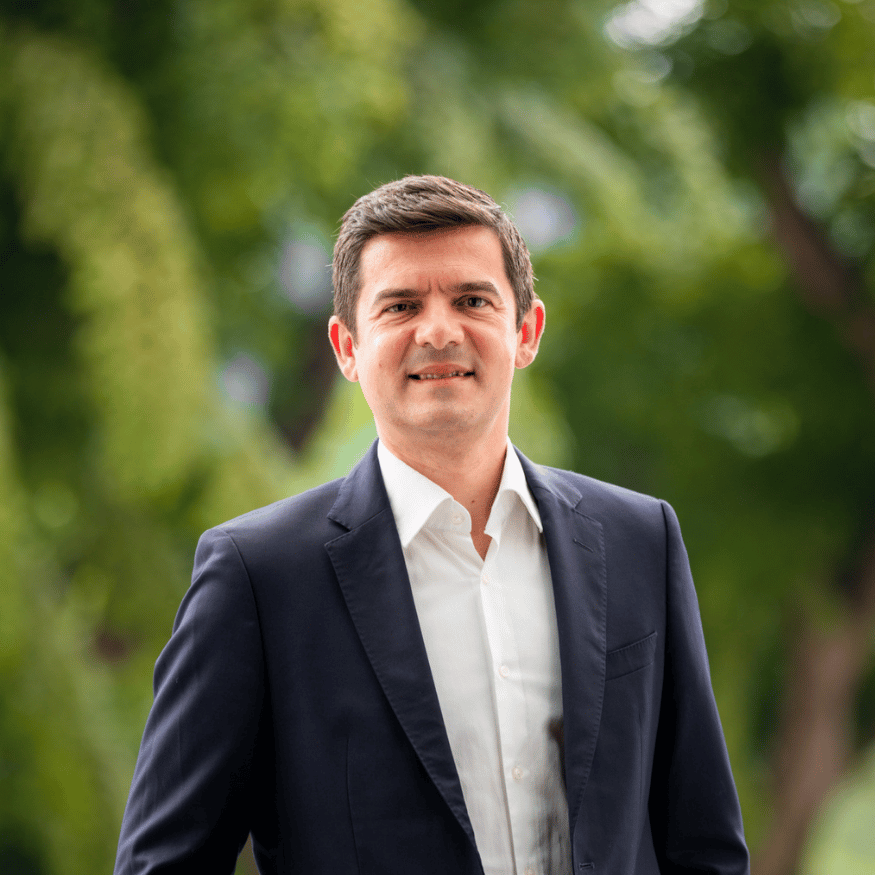 Picture of Andre Fernandes in a dark blazer standing outdoors with green trees in the background
