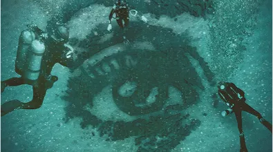 underwater art installation by artist alexandre farto aka vhils featuring a large eye carved into the seabed, surrounded by scuba divers exploring the intricate details of the artwork