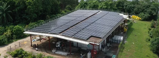 a large industrial building with solar panels covering its roof, located near a green field and a body of water. surrounding the building are various industrial materials and equipmen