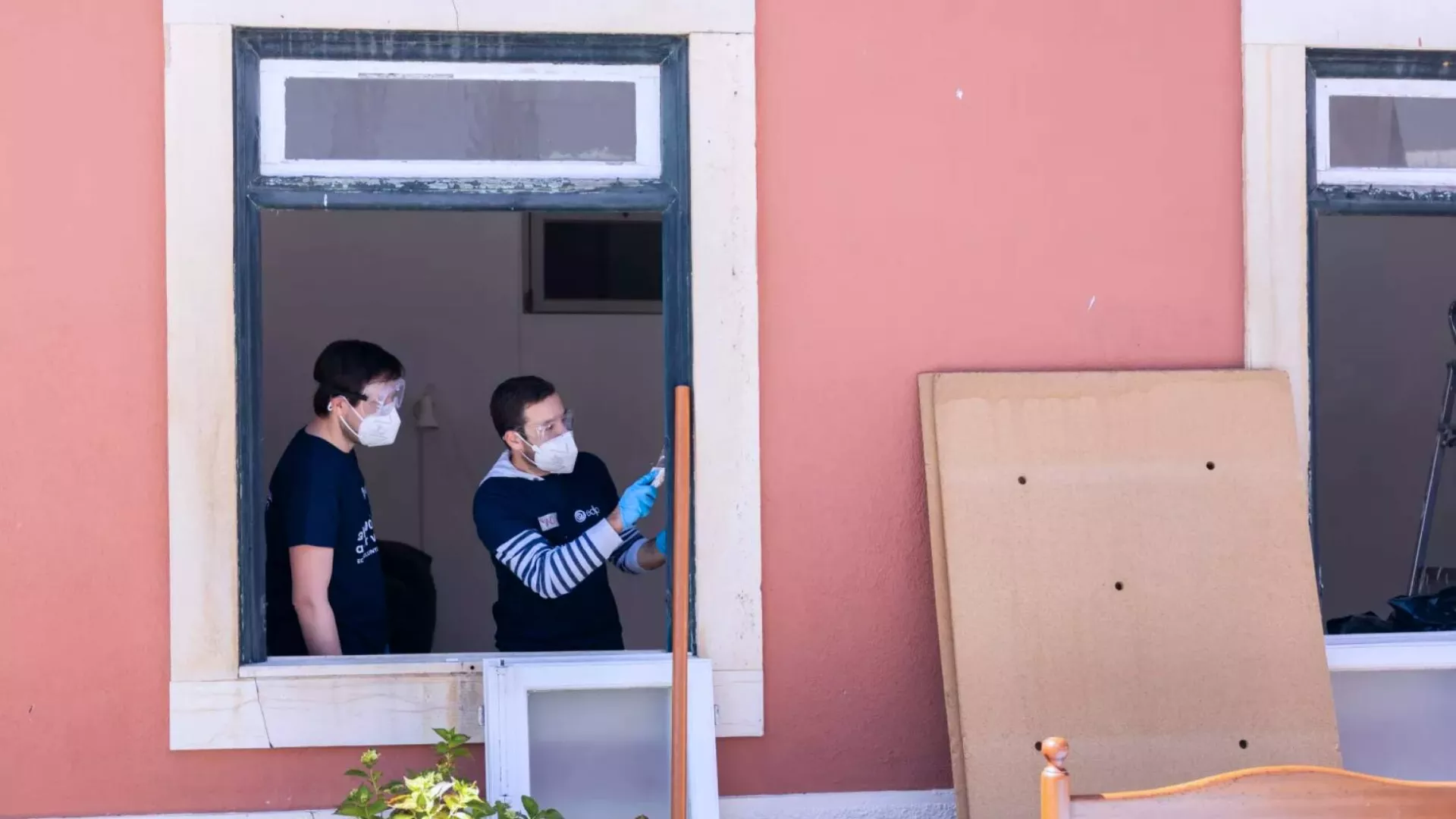two volunteers working on a  project, seen through a window. one person is painting a window frame with a brush, wearing gloves and a mask, while another observes or assists. outside, a piece of cardboard leans against a wall, suggesting renovation or cleanup efforts