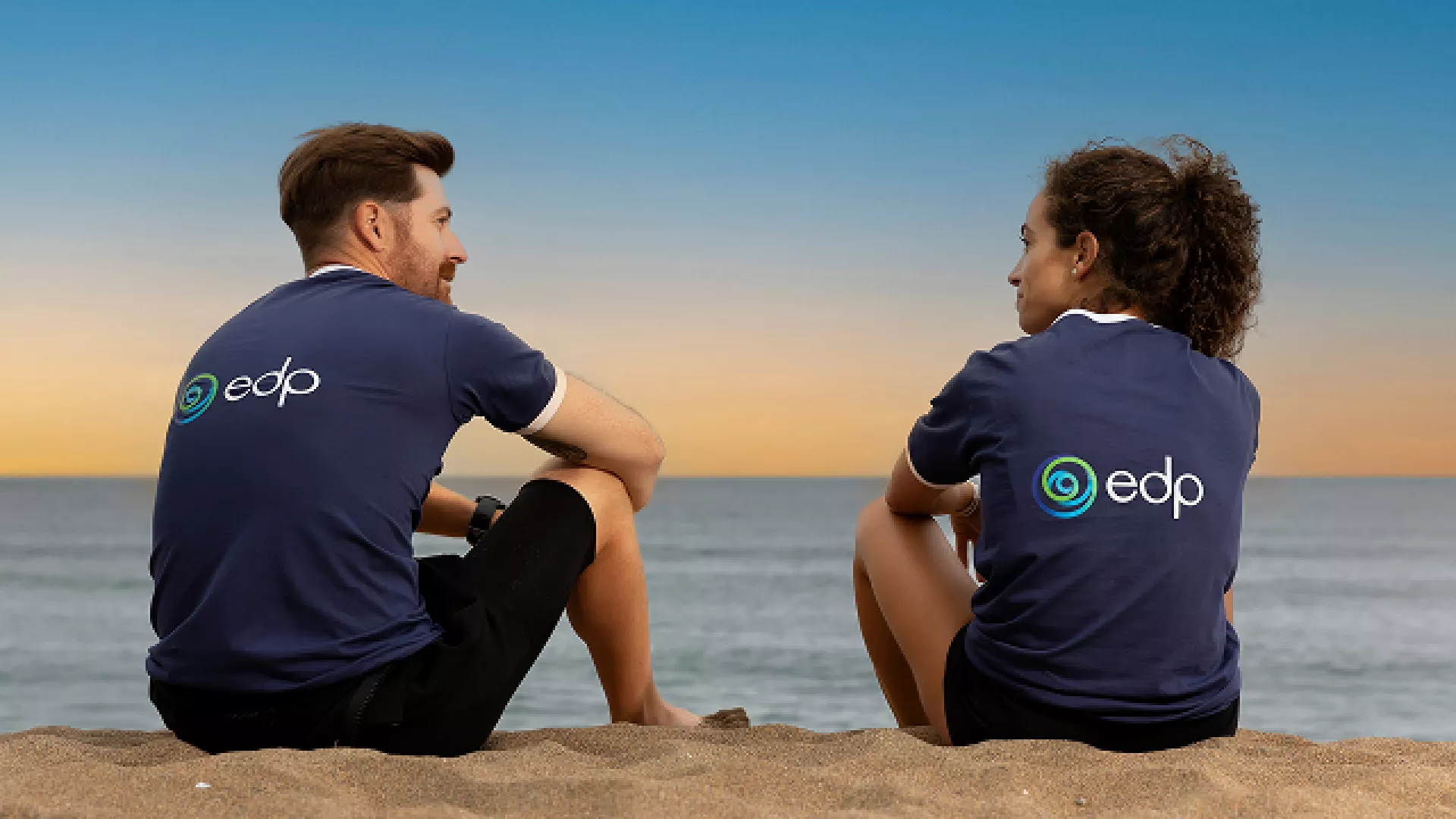 two young adults sit on the beach contemplating the sea, wearing edp tshirts