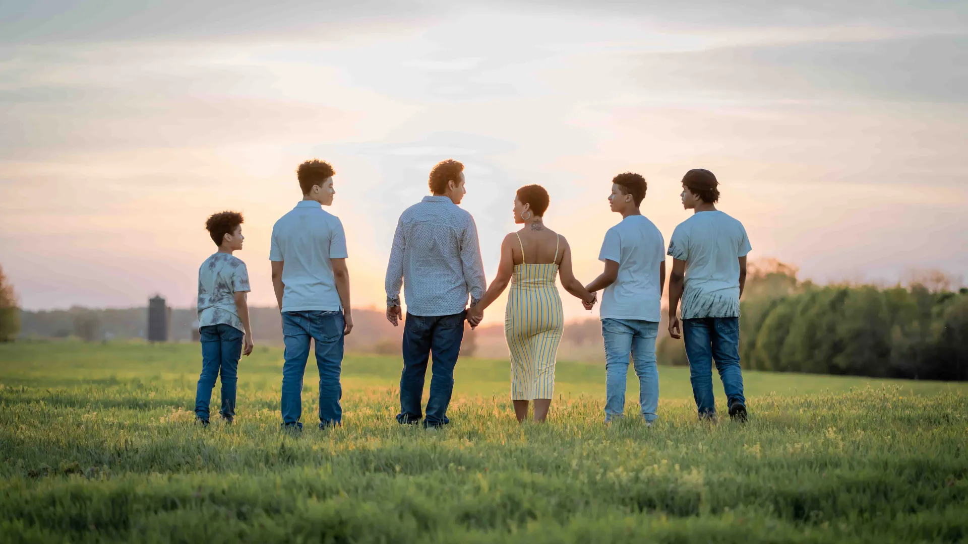 Family of six standing in a green field at sunset, holding hands and facing away