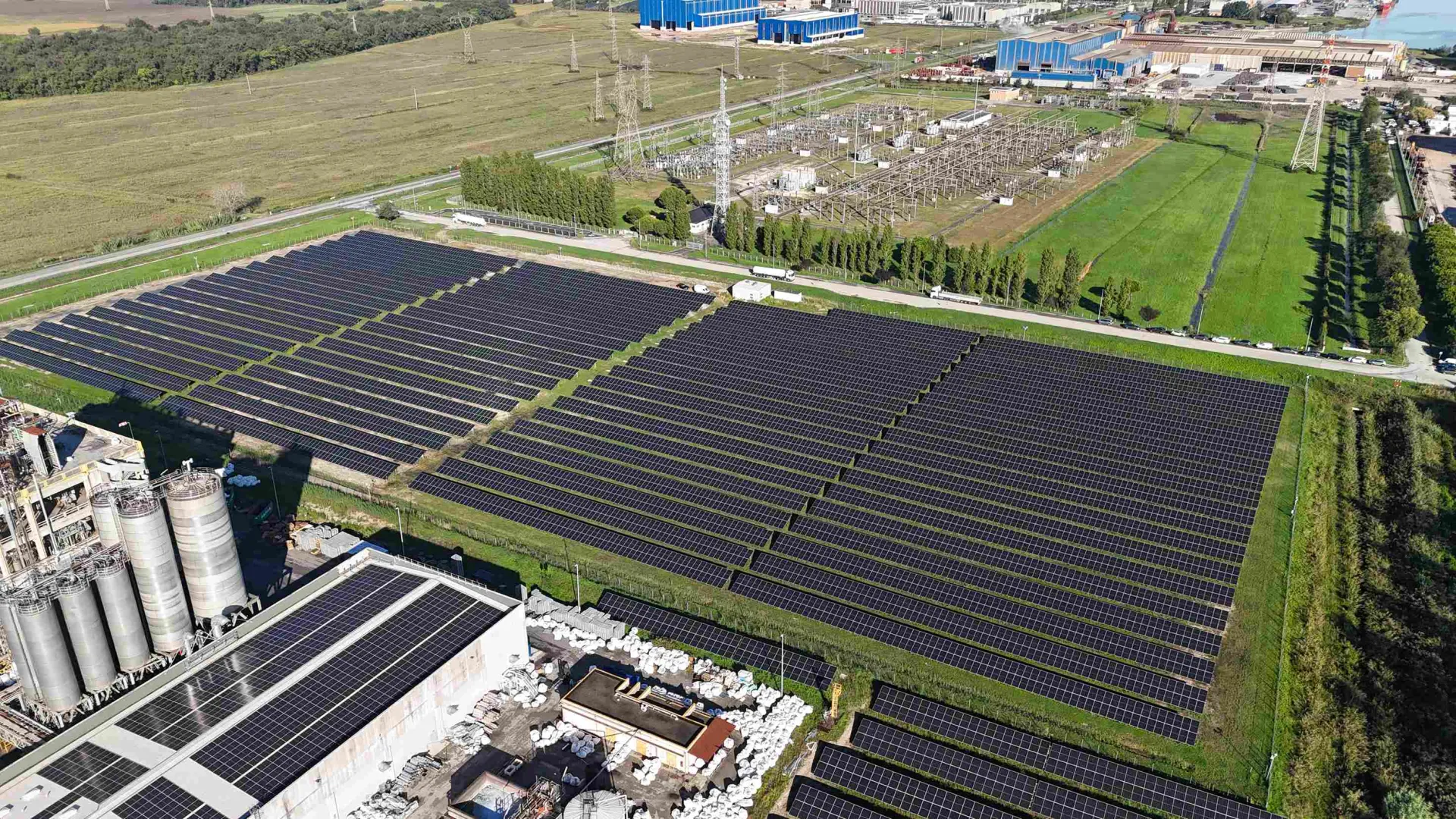 Aerial view of large solar panel fields beside industrial buildings and green farmland