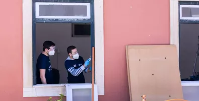 two volunteers working on a  project, seen through a window. one person is painting a window frame with a brush, wearing gloves and a mask, while another observes or assists. outside, a piece of cardboard leans against a wall, suggesting renovation or cleanup efforts