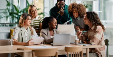 A group of people gathered around a laptop in a modern office, collaborating on a project and reviewing documents together.